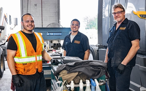 Three Schneider diesel technicians stand around a toolbox between service bays that are occupied by company trucks.