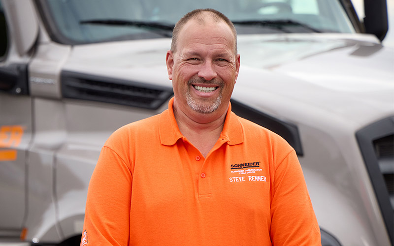 Steven Renner standing outside in front of a gray Schneider semi-truck, smiling straight ahead.