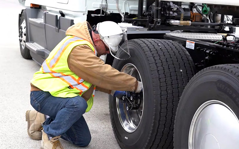 A truck driver in a hi-vis vest and a hard hat inspecting one of the wheels on their semi-truck.