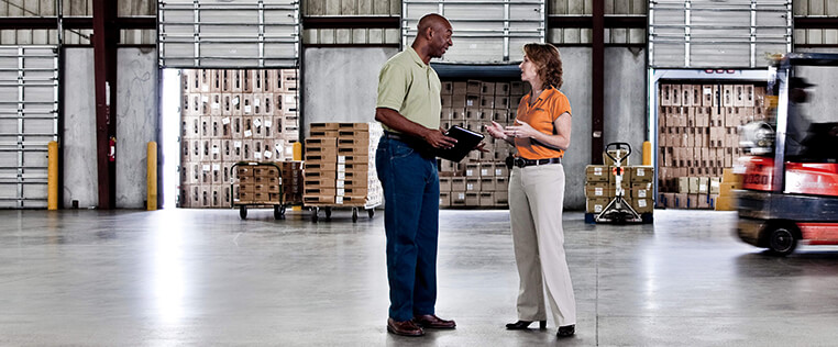 Two Schneider associates speak in a company warehouse with a forklift passing by