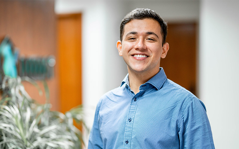 A man wearing a blue button up shirt standing and smiling inside an office.