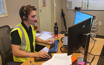 A young man wearing a safety vest and headset sits at his desk with his hands on his keyboard.