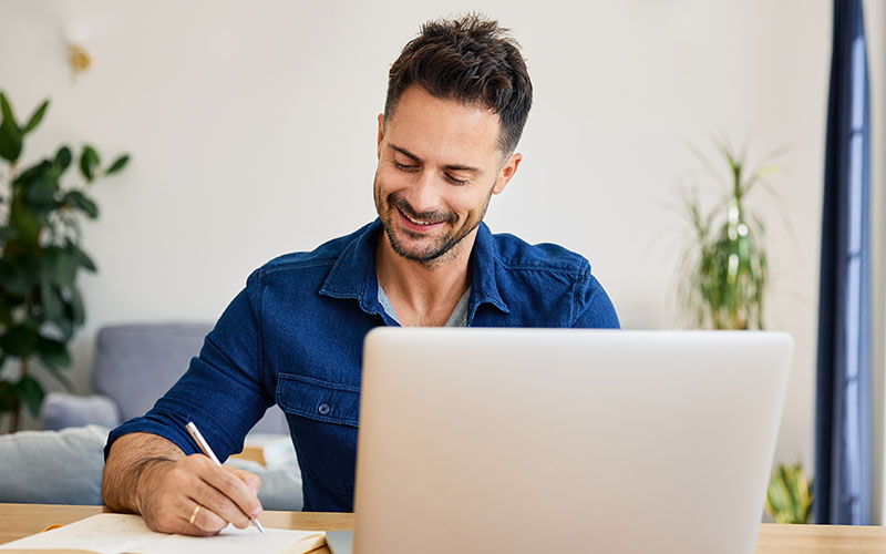 A man sitting a table and writing on a piece of paper with an open laptop in front of him.