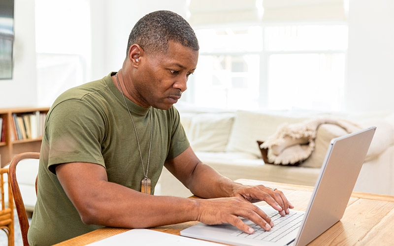 A man in a green shirt and dog tag in his living room looking at his laptop. 