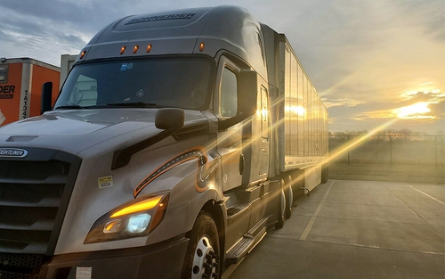 A grey Schneider semi-truck is parked in a lot with a golden sunset in the background.