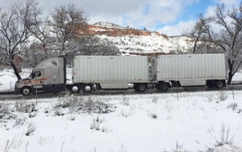 A grey Schneider semi-truck hauling two white trailers is parked at a rest area. There is snow on the ground a snow-covered mountain and trees in the background.