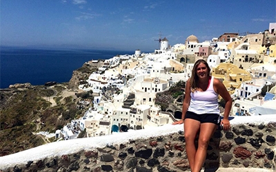 Samantha May leans against a stone wall overlooking a Greek village during her travels.