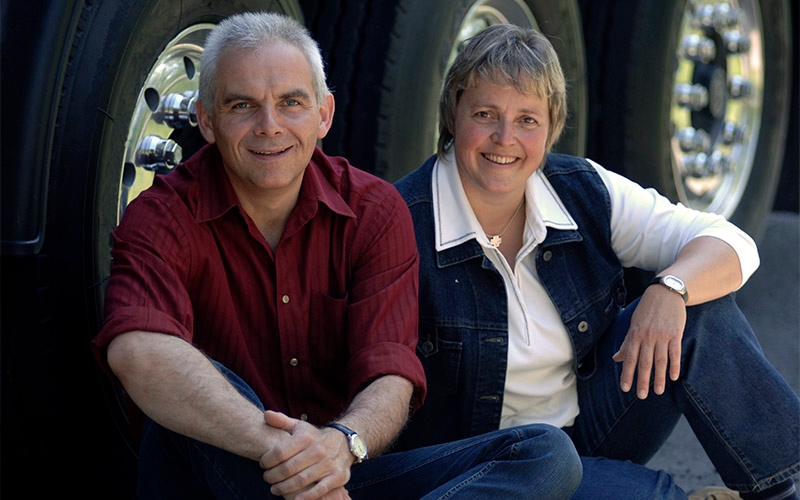 A man and a woman sit on the ground in front of the tires of an eighteen wheeler.