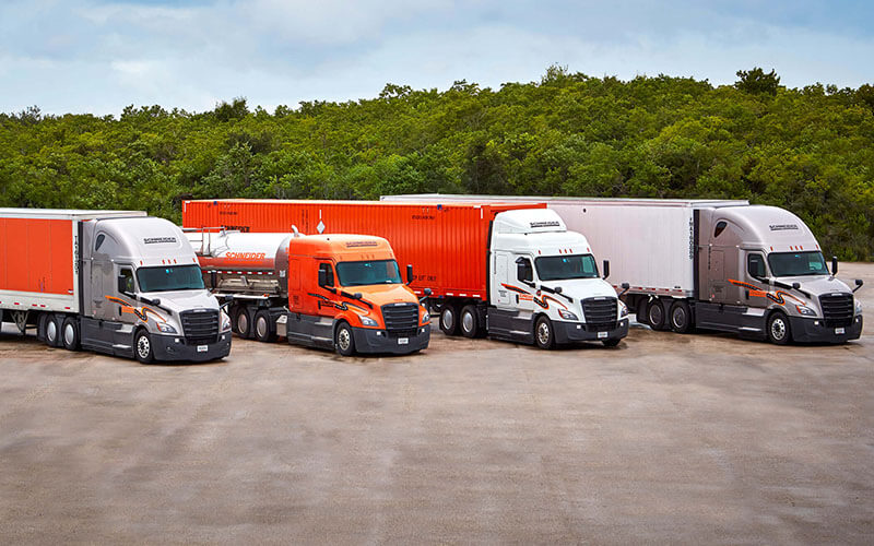 Four Schneider semi-trucks are parked at an angle in a parking lot. Each truck is hauling a different trailer, representing each of Schneider's lines of service.