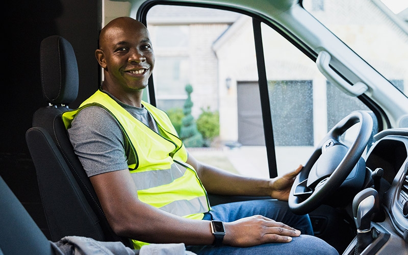 A professional driver wearing a high-visibility vest seated in the driver’s seat of a vehicle, hands resting on the steering wheel.