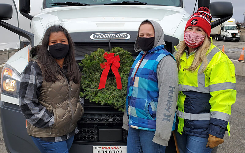 Three women who are wearing masks and dressed for cold weather, stand in front of a white Freightliner Cascadia semi-truck that has a wreath on it.