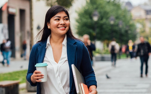 A female associate smiles brightly while walking down a busy pathway with a laptop and notebook tucked under one arm and a to-go coffee cup in hand.