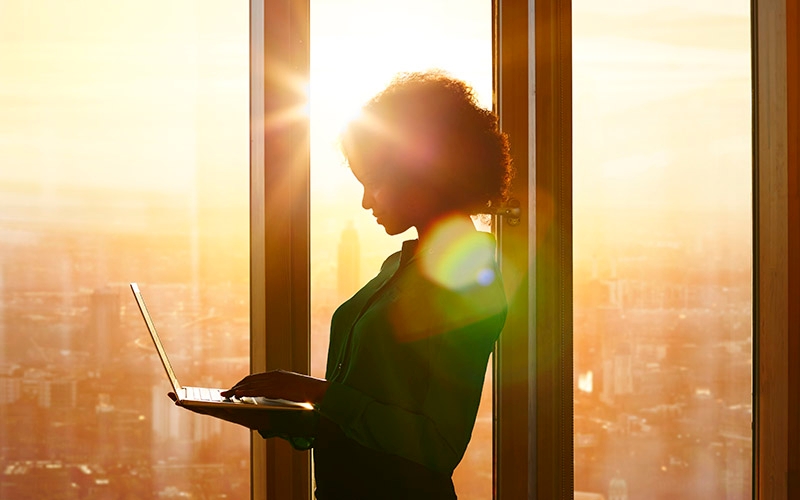 A woman standing in front of a large window with the sun rising on the horizon. She is holding a laptop and looking down at the screen.