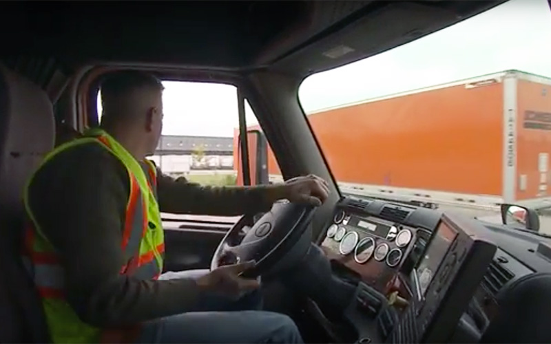 A truck driver wearing a neon safety vest drives past an orange Schneider trailer.