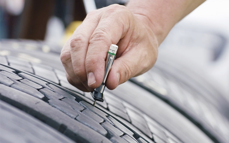 A close-up view of a hand using a tire tread depth gauge to measure the tread depth on a semi-truck tire. 