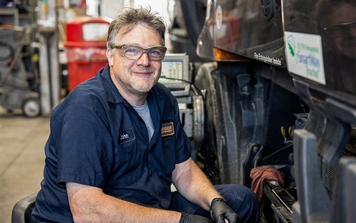 A man sitting beside a semi-truck wearing safety glasses and smiling.