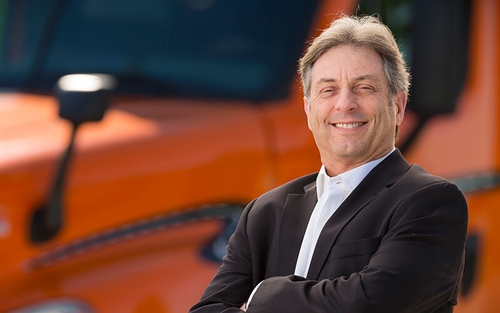 Max Pietsch smiles and stands in front of an orange Schneider semi-truck with his arms crossed.