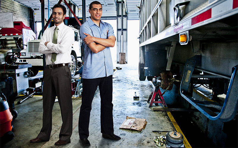 Two men standing with their arms crossed in a diesel mechanic shop next to a semi trailer. One is wearing a white dress shirt, tie, black dress pants and dress shoes and the other is wearing a blue dress shirt, black dress pants and dress shoes.