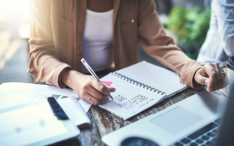 A close up shot shows a woman wearing a white shirt and tan jacket and writing in a notebook with a laptop in front of her and notebooks to the right of her.