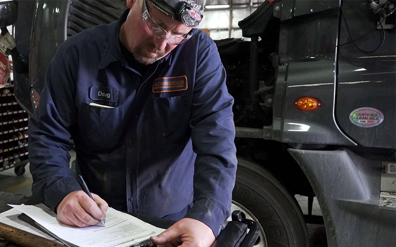 A Schneider diesel technician in a shop filling out paperwork on a clipboard. A semi-truck is parked behind him.