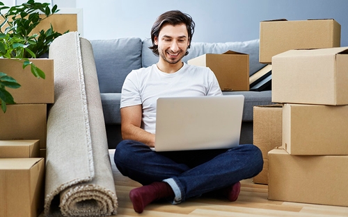 A man sitting on the floor with cardboard boxes around him and looking at a computer 