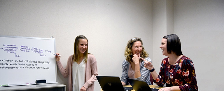 A Schneider associate gestures towards a whiteboard with financial information written on it while two other colleagues smile and converse with her 