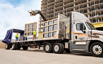 A grey semi-truck with a Conestoga trailer attached to it is unloaded by construction workers.