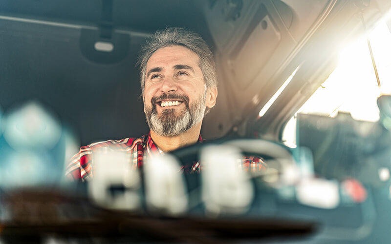 A truck driver sitting behind the wheel of a semi-truck smiles as he drives.