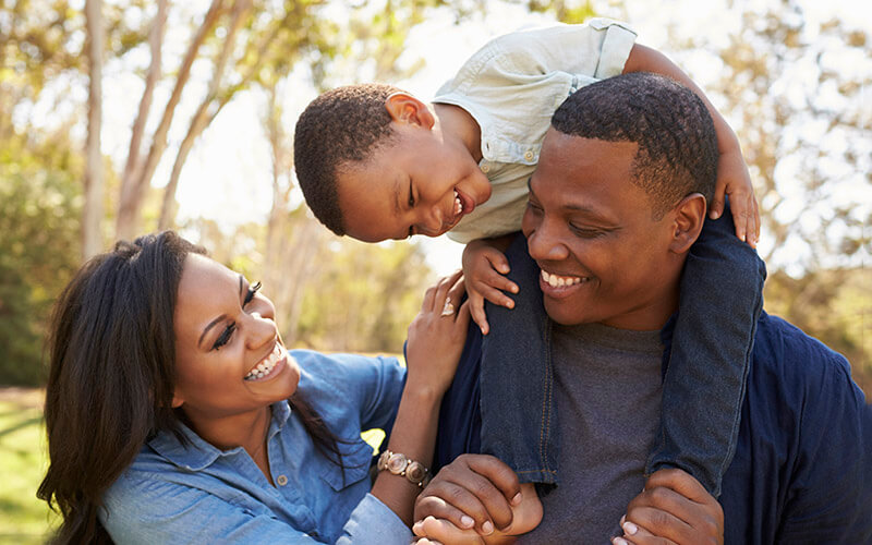 An African-American family smiles and laughs as they play together outside. A little boy sits on his father's shoulders and laughs as his mother tickles him.