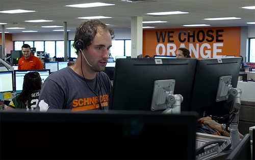 A man wears a headset and types on a set of two monitors within an office setting. A wall behind him has the words "We chose orange" on it.