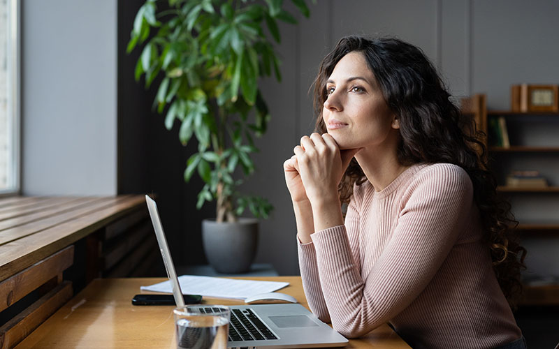 A woman sitting at a desk staring out the window with a hopeful expression on her face.
