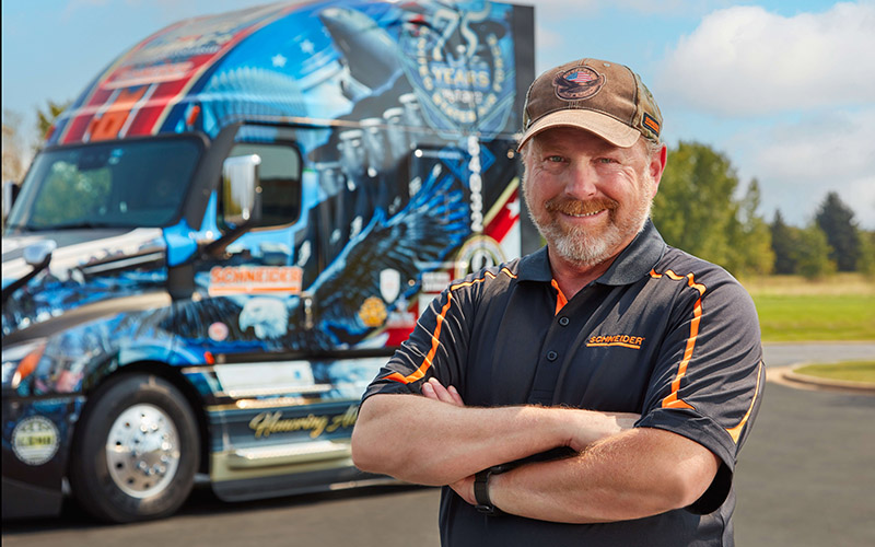 Schneider 2022 Ride of Pride driver, Jeff Waggoner, stands in front of his Ride of Pride truck with his arms crossed.