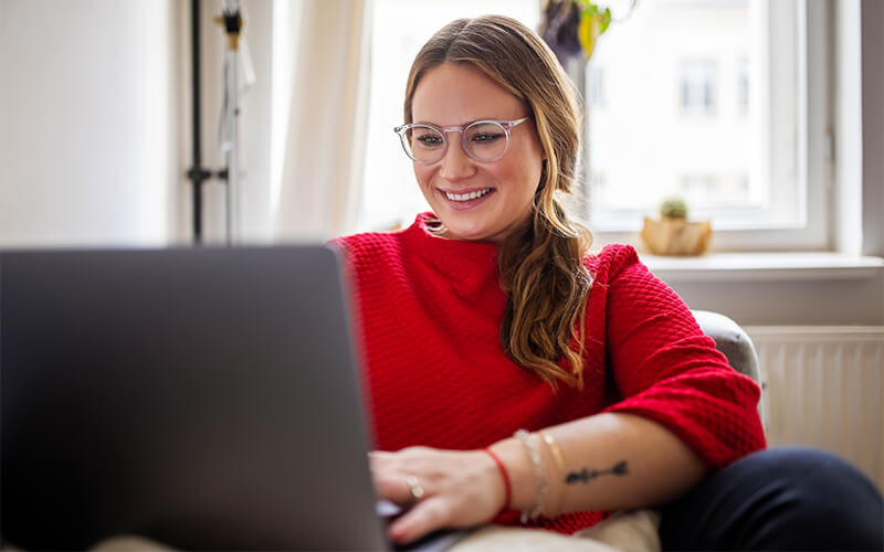 A lady with a red sweater and blue light glasses sits on a couch and works on a laptop.