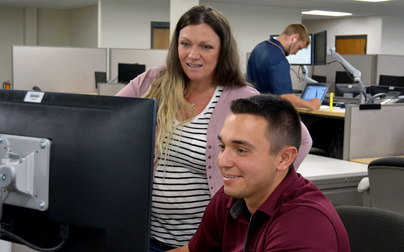 In an office setting, a woman stands next to a man who is sitting down and looking at his computer.