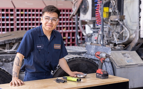 A Schneider diesel technician, wearing a blue jumpsuit and a name tag titled, 'Chavez,' stands in front of a work table in a shop with tools on top of it.