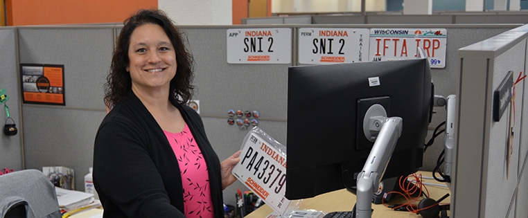 A Schneider Purchasing associate stands at her desk holding a new license plate for a Schneider truck while reviewing documents on her computer.