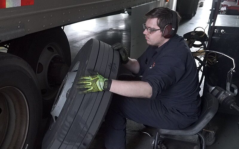 A male diesel technician putting a tire on a semi-truck. He's wearing safety glasses, gloves and ear protection.