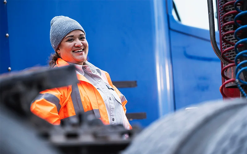 A truck driver in an orange safety vest and grey hat standing in front of a blue semi-truck cab. 
