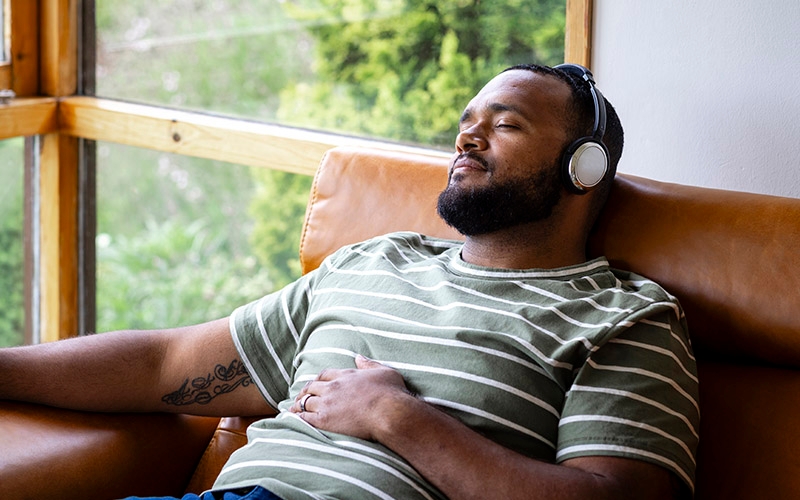 A person wearing headphones and a green striped shirt reclining on a brown leather couch near a window with greenery outside.