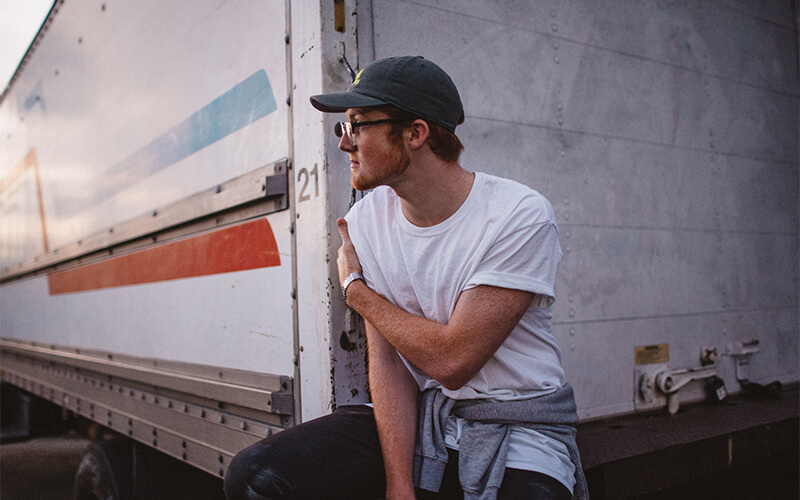 A man in a white t-shirt and grey hat sits on the edge of a white semi-trailer.