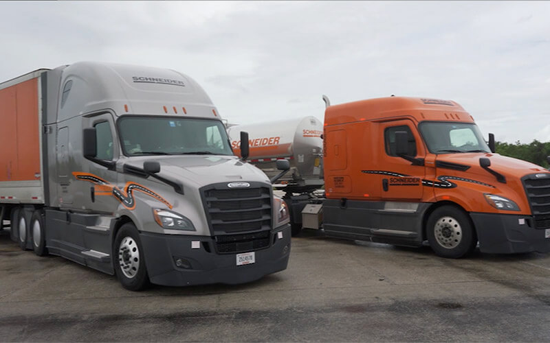 Two Schneider trucks, a grey truck with an orange trailer and an orange truck with a tanker trailer, are parked at an angle in a parking lot.