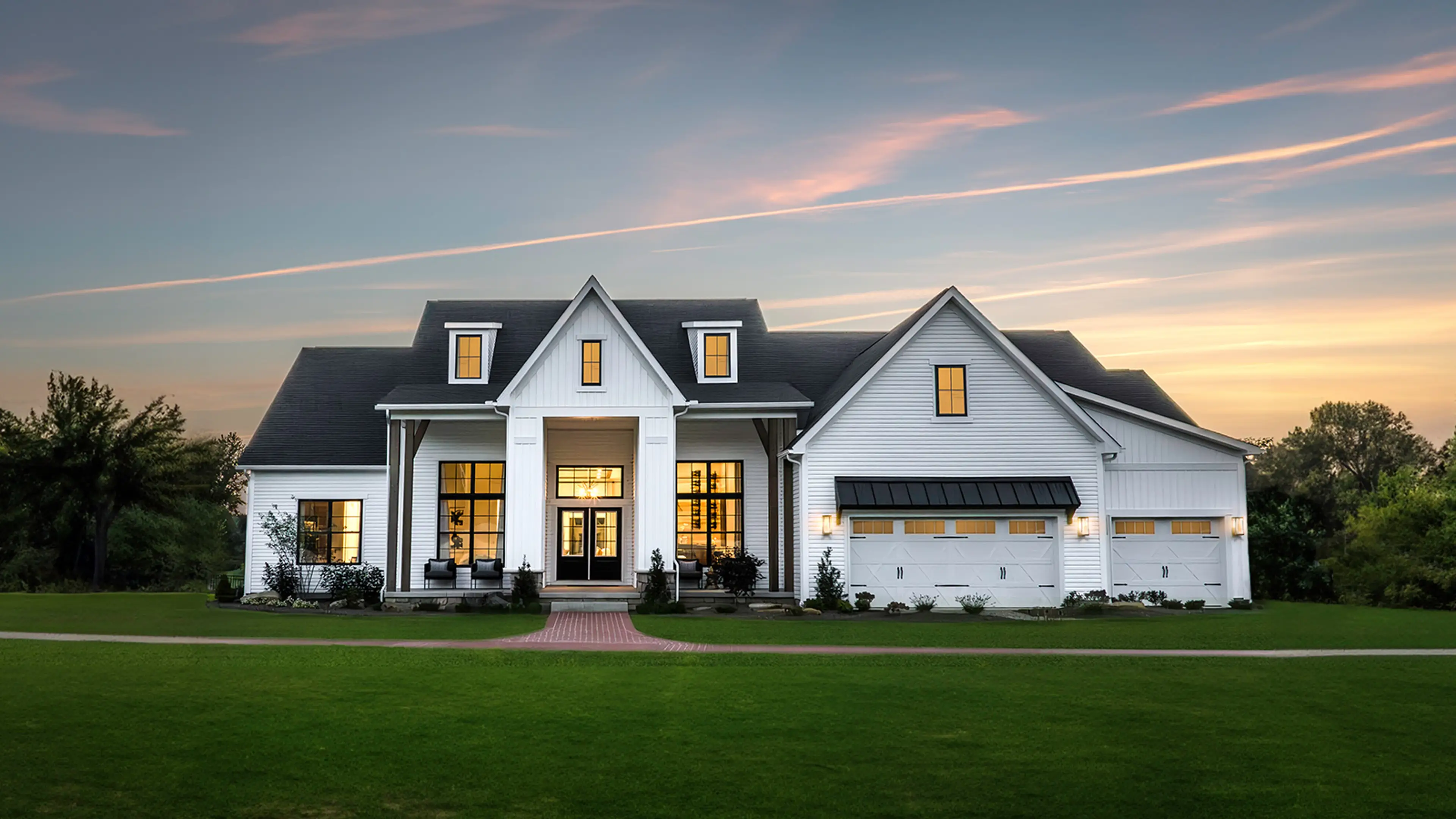 Exterior view of the Charleston model home, a large white farmhouse with a black roof and a brick walkway. The sunset casts a golden glow on the lawn and lights the interior.
