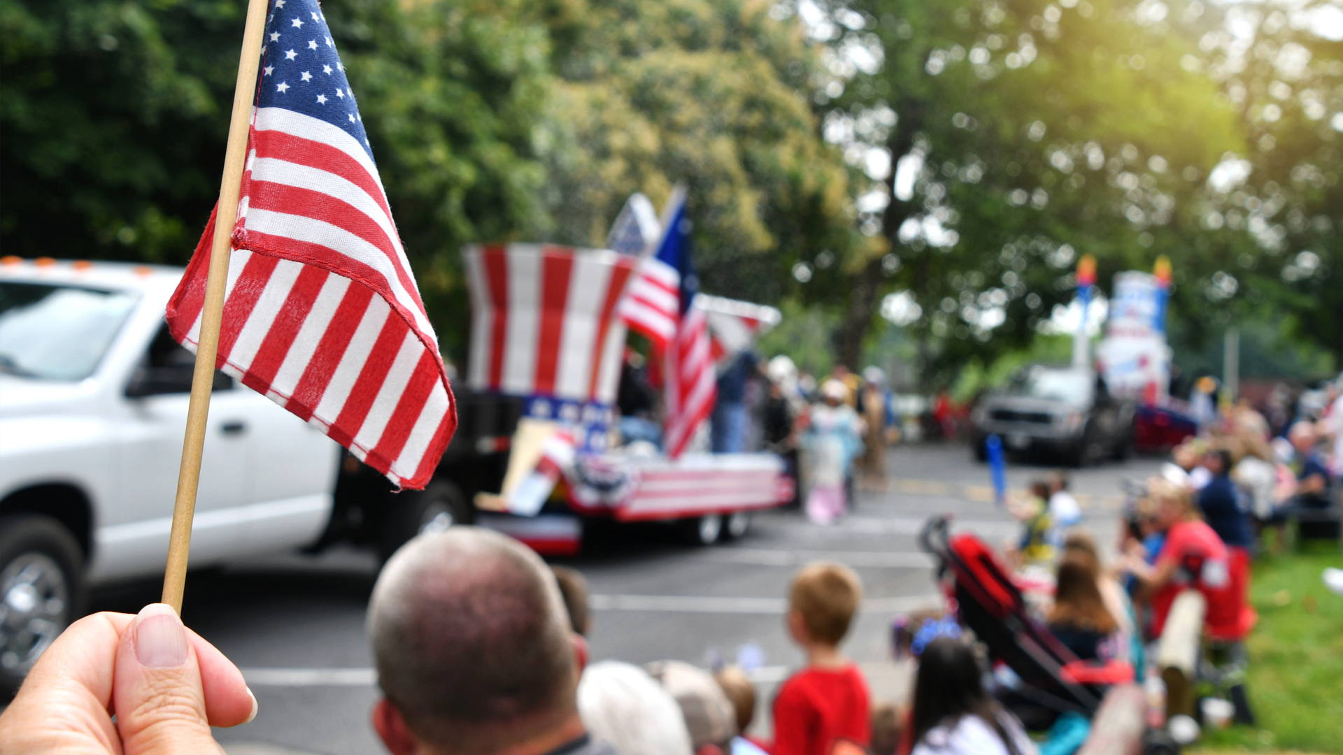 Community parade scene with people gathered along a street and multiple American flags displayed on vehicles and throughout the crowd
