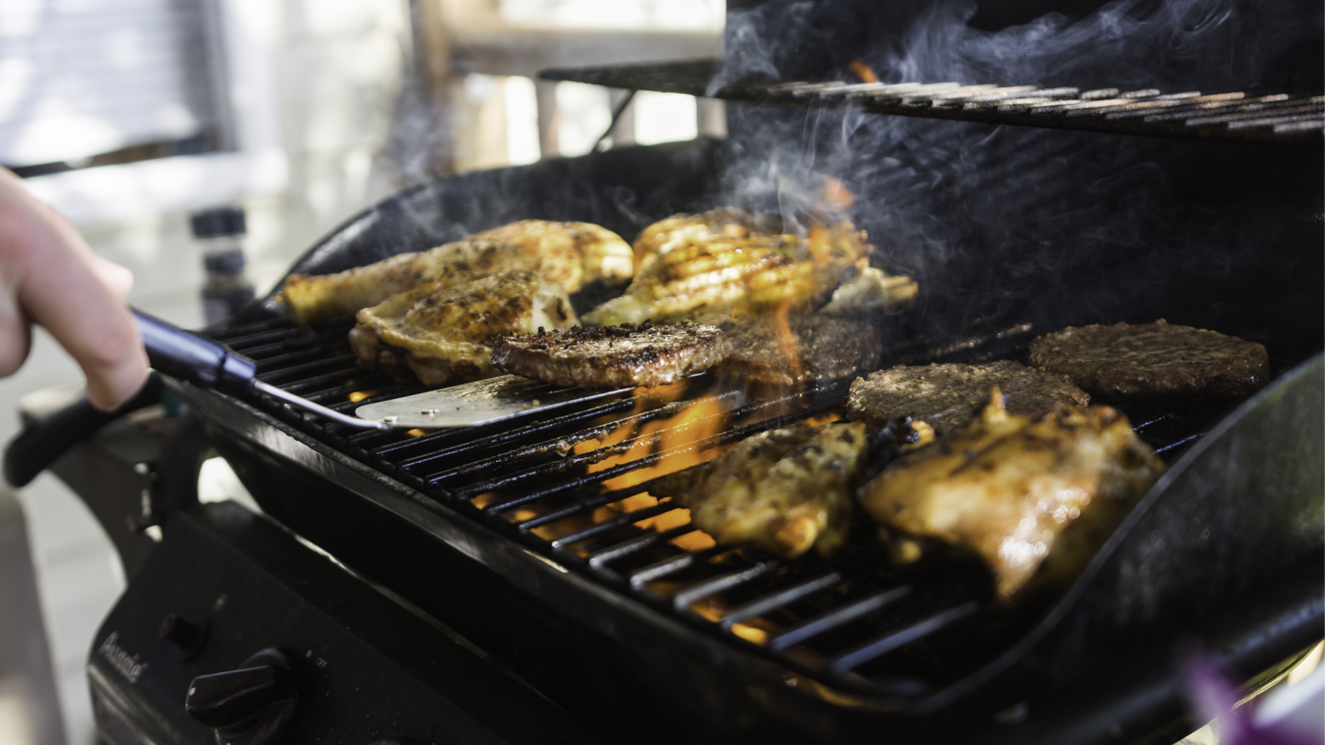 Close-up of chicken and burgers cooking on a gas grill, with smoke and flames rising as a person turns the food with a spatula.