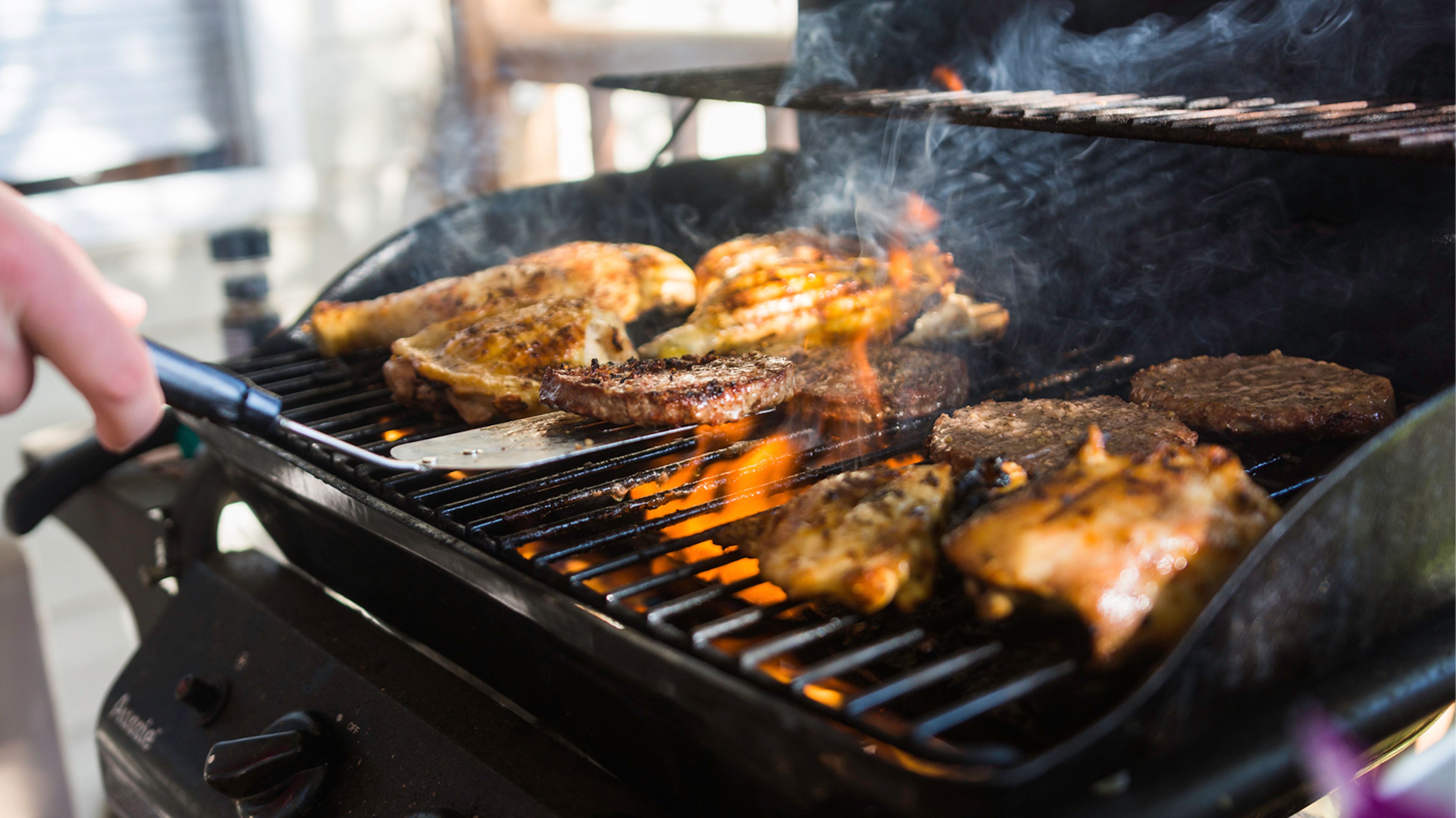 Close-up of chicken and burgers cooking on a gas grill, with smoke and flames rising as a person turns the food with a spatula.