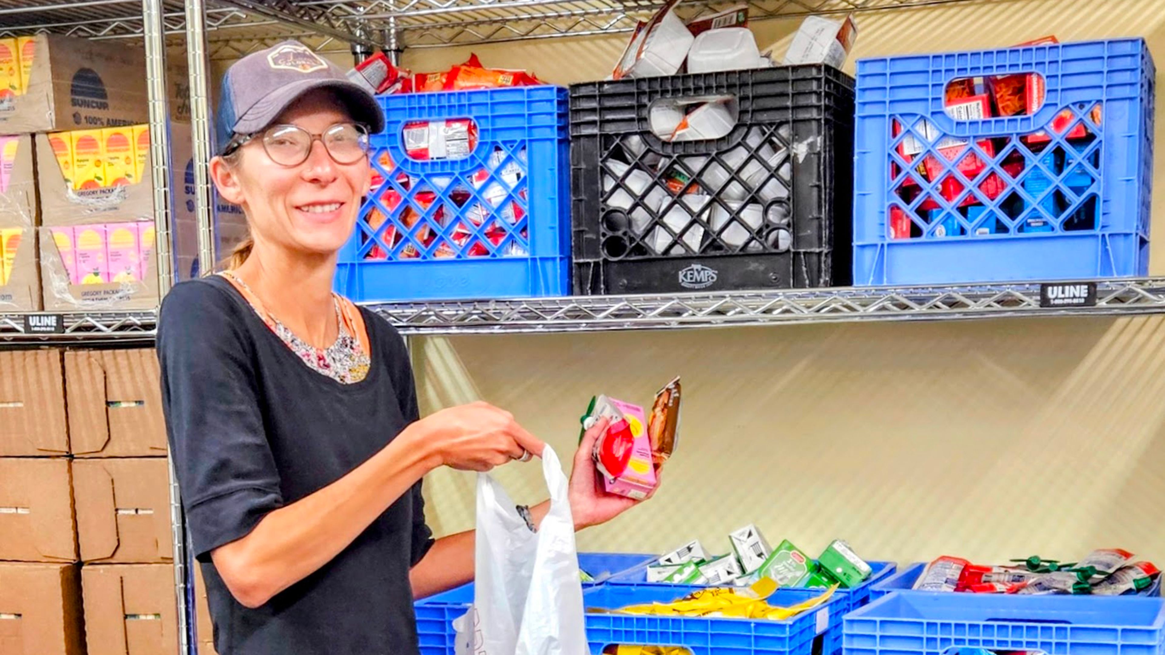 Smiling woman in cap and glasses holding reusable bag and packaged food inside a food-distribution area. Blue and black plastic crates, shelves, boxes, and assorted produce or packaged goods create a warehouse-style setting.