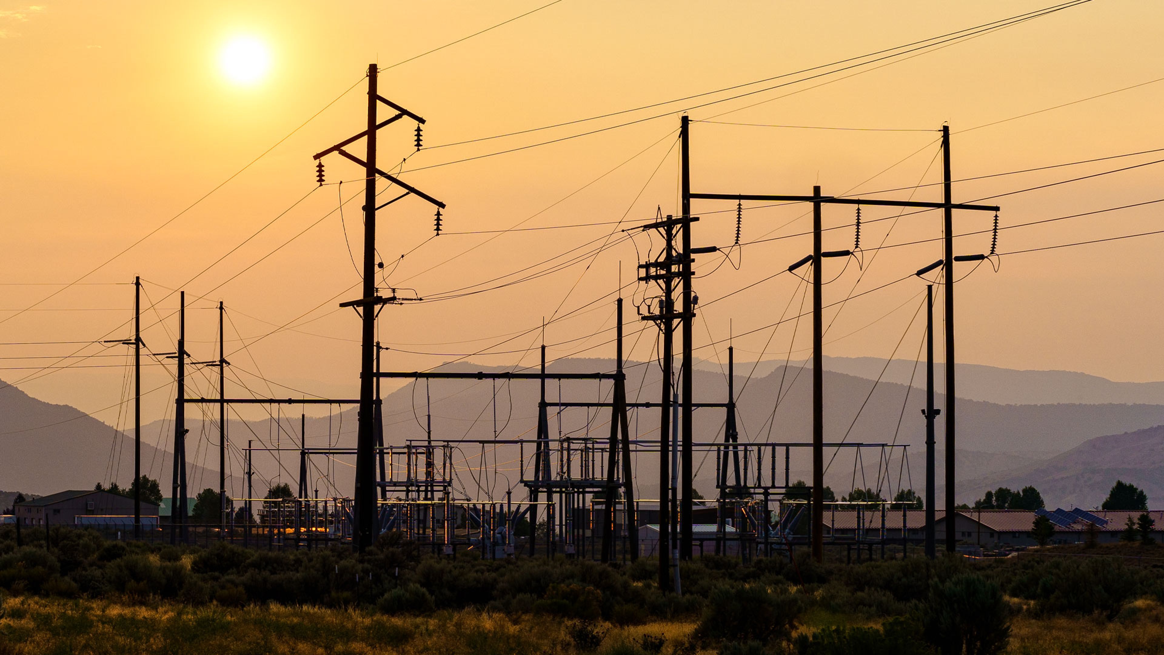 Electrical substation with multiple power lines and tall transmission towers silhouetted against a hazy sunset sky, with mountains in the background and vegetation in the foreground.