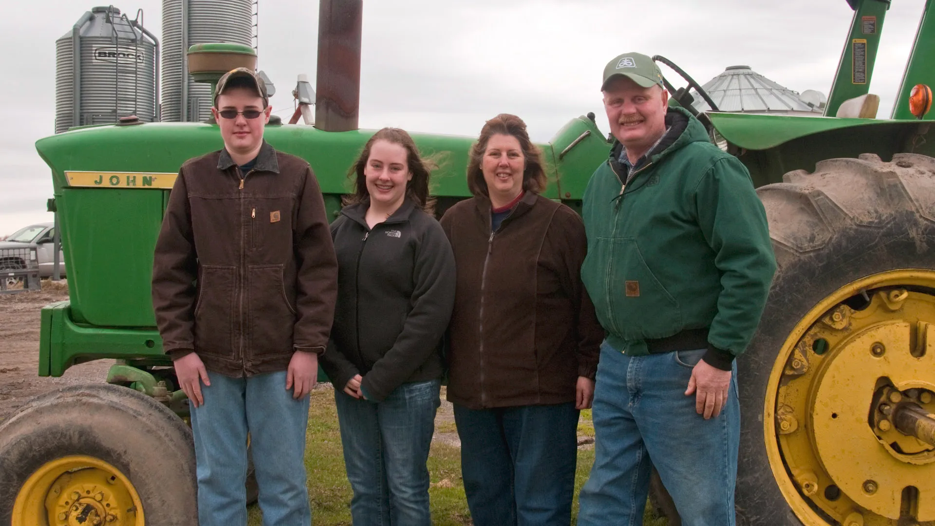 Four people standing in front of a large green farm tractor with grain silos in the background.
