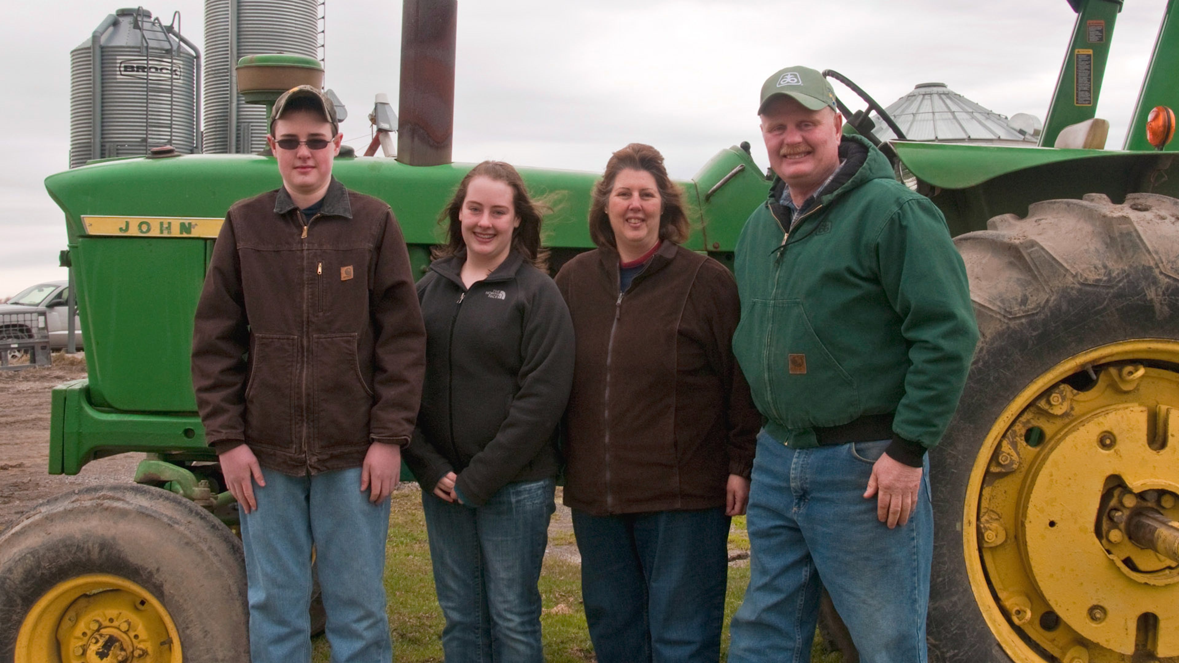 Four people standing in front of a large green farm tractor with grain silos in the background.