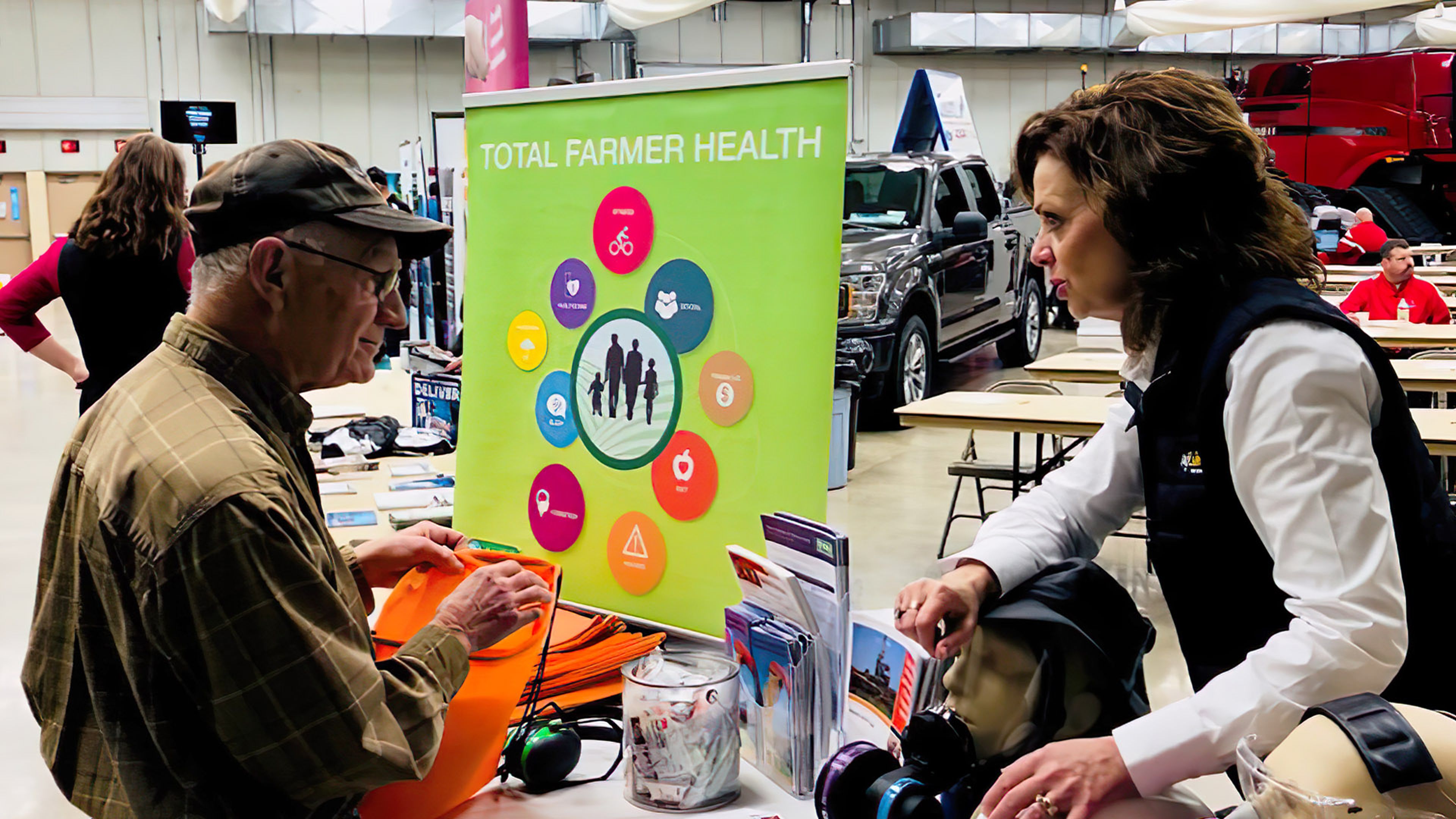 Two people speaking at a booth featuring a “Total Farmer Health” display with brochures and safety gear.